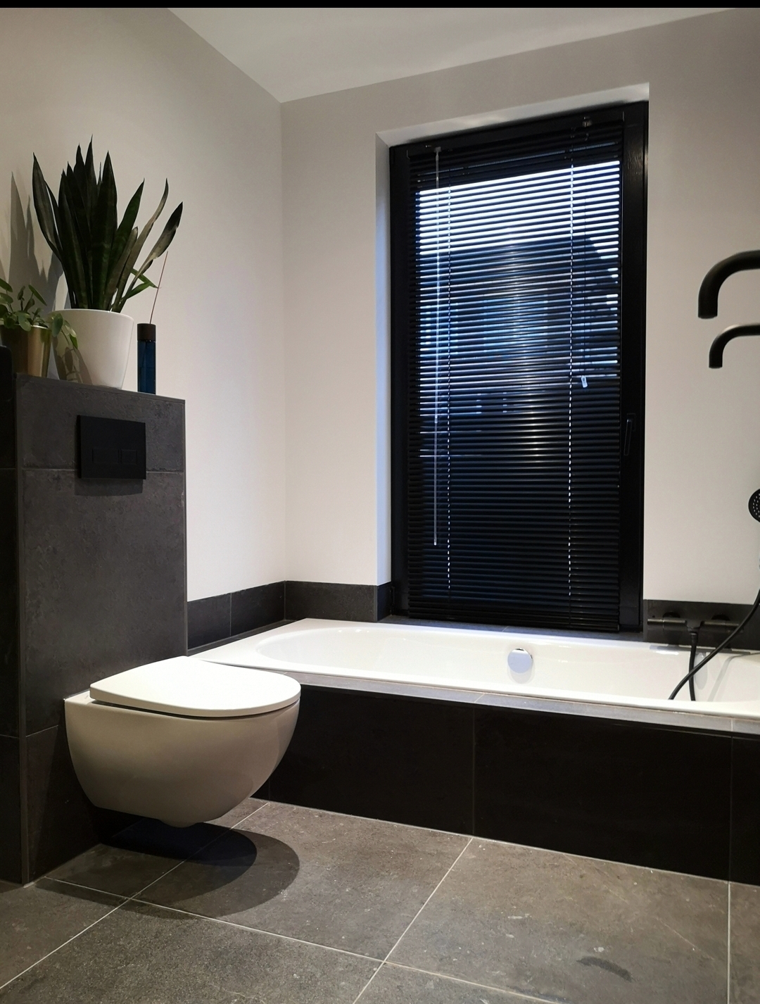 Door KeraPro gerenoveerde badkamer: donkere natuursteenvloer, ligbad onder het raam en een hangend toilet